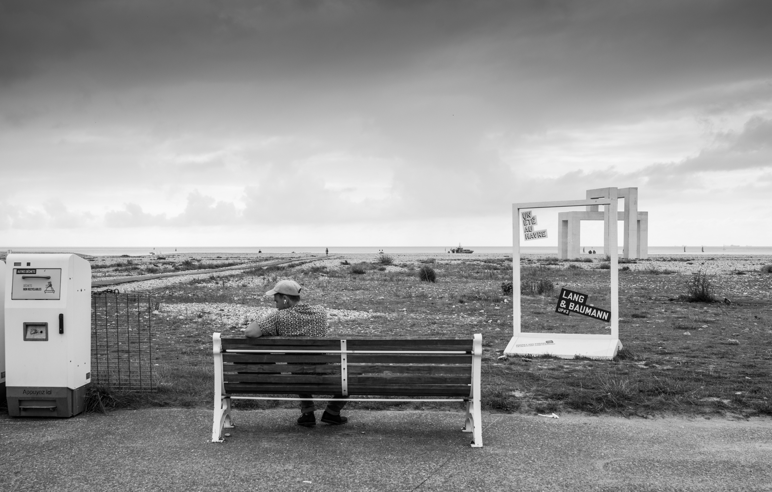 Ein Sommer in Le Havre. Ein Mann sitzt vor dem eingerahmten Horizont des bleiweißen Meeres. Schräg vor ihm Rahmen, durch die man auf den entfernten Horizont schauen kann.